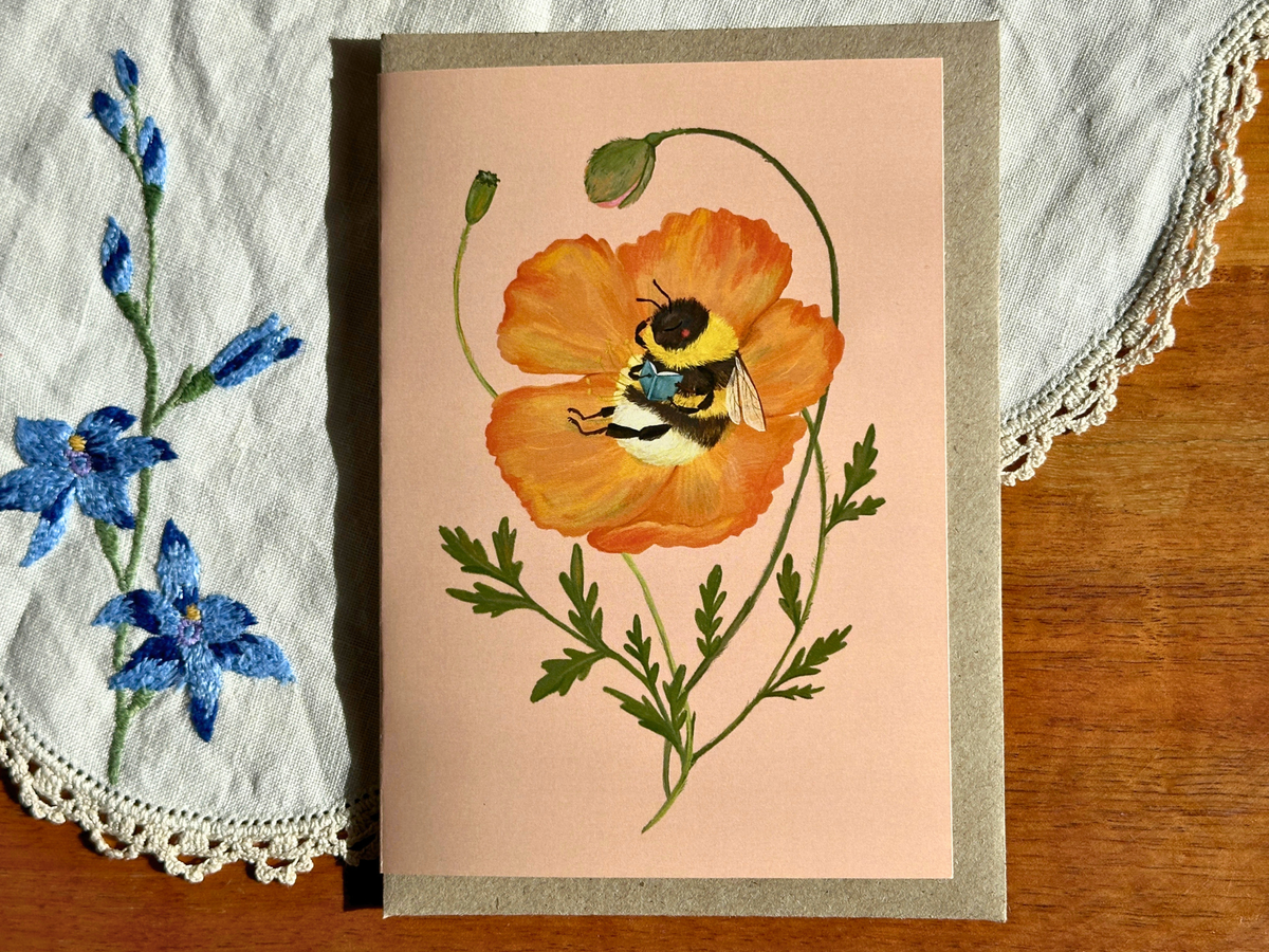 Beebliophile greeting card with a reading bee in an orange poppy, on an embroidered doily on a wooden table