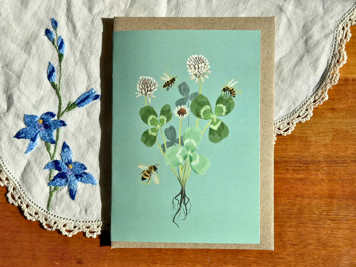 Clover greeting card with clover and honeybees on mint green, laid on an embroidered doily on a wooden table