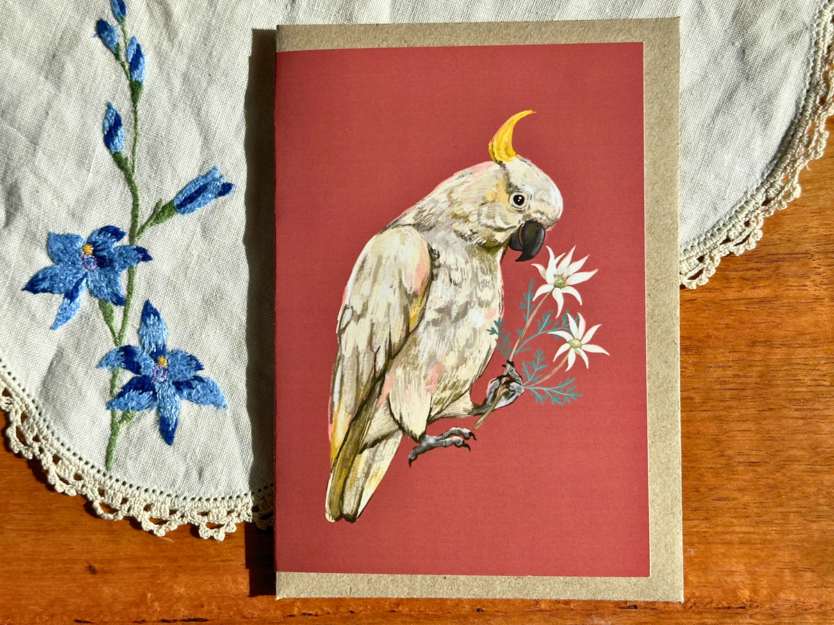 Cockatoo greeting card with a cockatoo and flannel flowers on terracotta, on an embroidered doily on a wooden table
