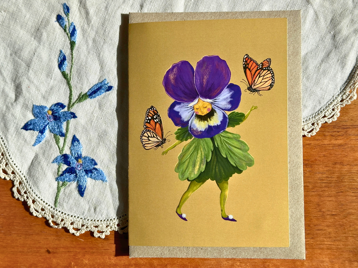 Pretty Pansy greeting card with a dancing pansy and butterflies, laid on an embroidered doily on a wooden table