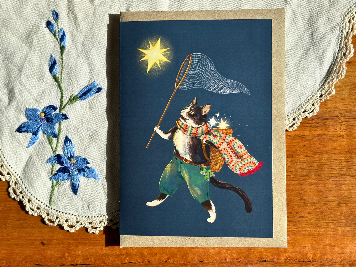 Ashy Cat greeting card with a star-catching cat on navy, laid on an embroidered doily on a wooden table