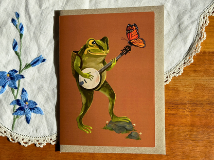 Banjo Frog greeting card with a frog strumming a banjo on burnt orange, on an embroidered doily on a wooden table