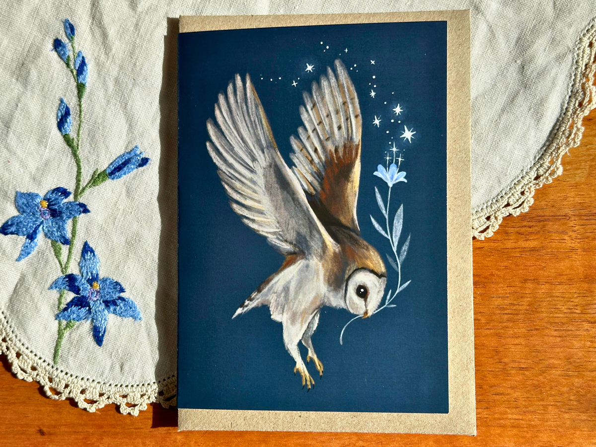Barn Owl greeting card with a swooping owl and starry blue flower, on an embroidered doily on a wooden table