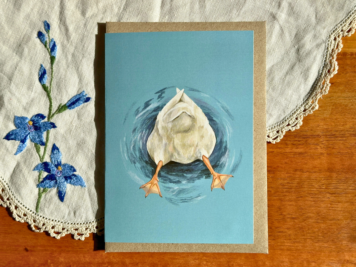 Butt Quack greeting card with a diving duck's bottom and feet on teal, on an embroidered doily on a wooden table