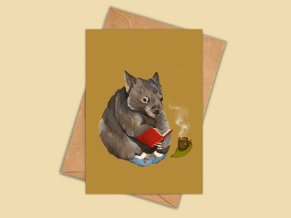 Cosy Wombat greeting card by Anna Seed: a wombat in slippers reading a red book beside a cup of tea, on mustard