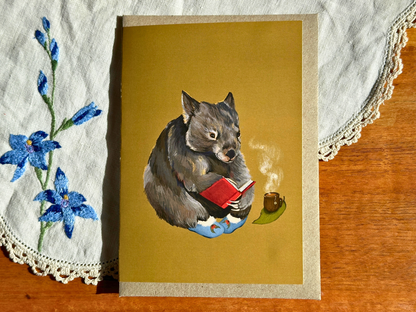 Cosy Wombat greeting card with a reading wombat and steaming tea, laid on an embroidered doily on a wooden table
