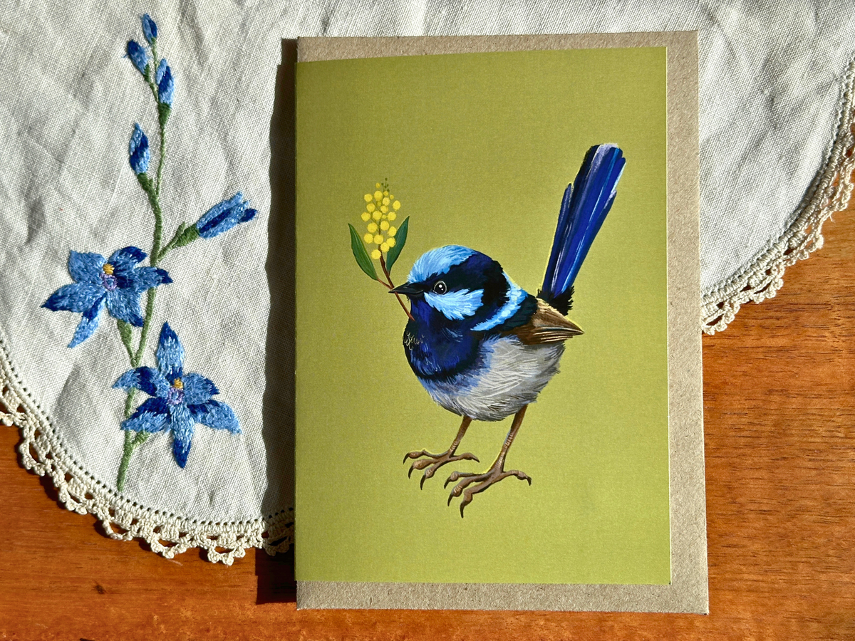 Fairy Wren greeting card with a blue wren and wattle sprig on olive green, on an embroidered doily on a wooden table