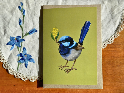 Fairy Wren greeting card with a blue wren and wattle sprig on olive green, on an embroidered doily on a wooden table