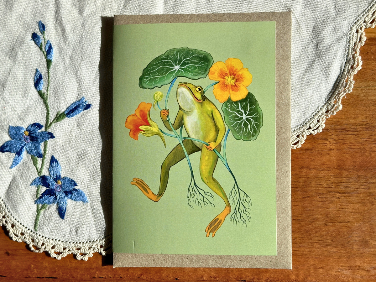 Happy Frog greeting card with a frog dancing among nasturtiums, laid on an embroidered doily on a wooden table