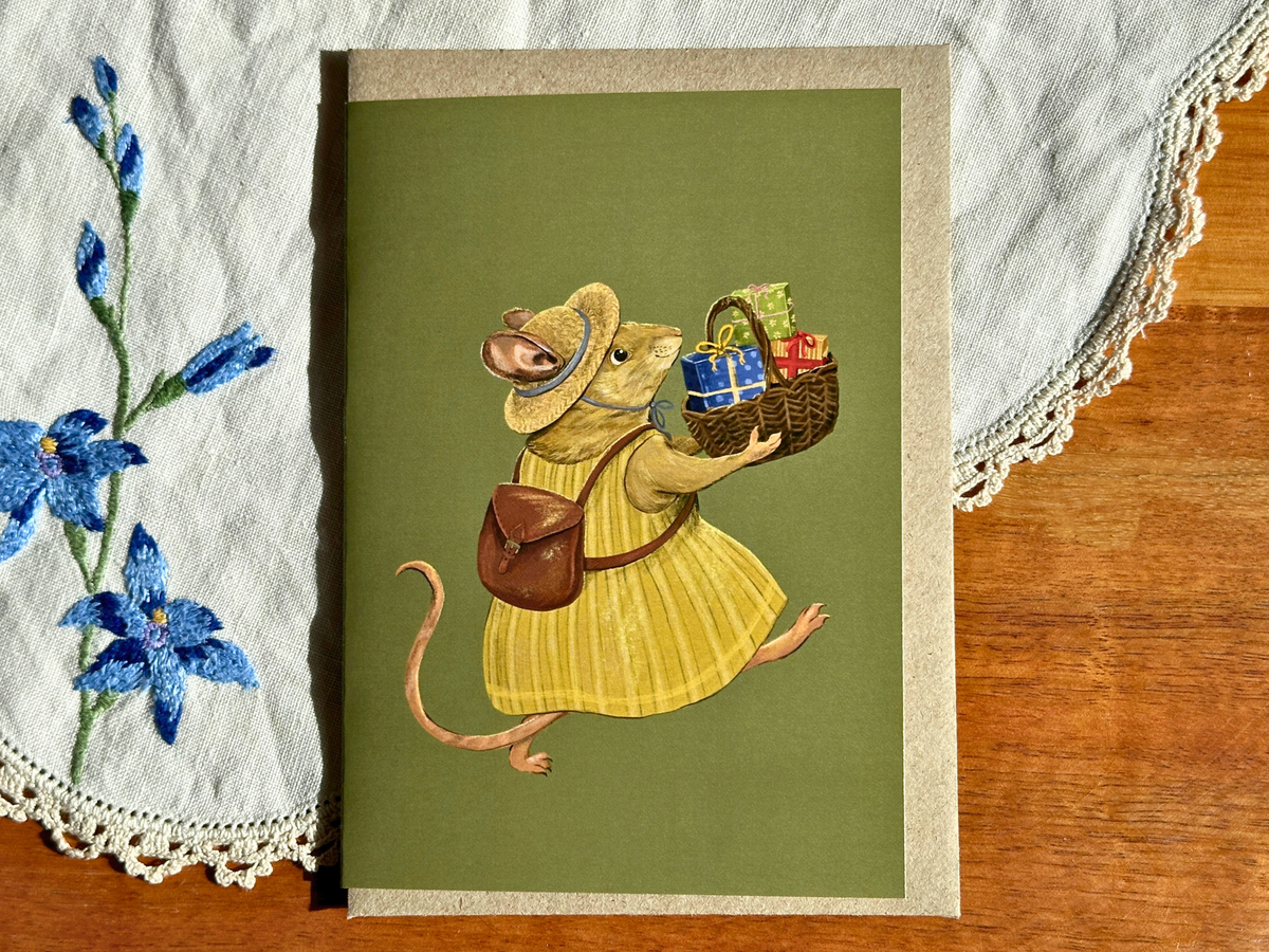 House Mouse greeting card with a mouse carrying a basket of presents, laid on an embroidered doily on a wooden table