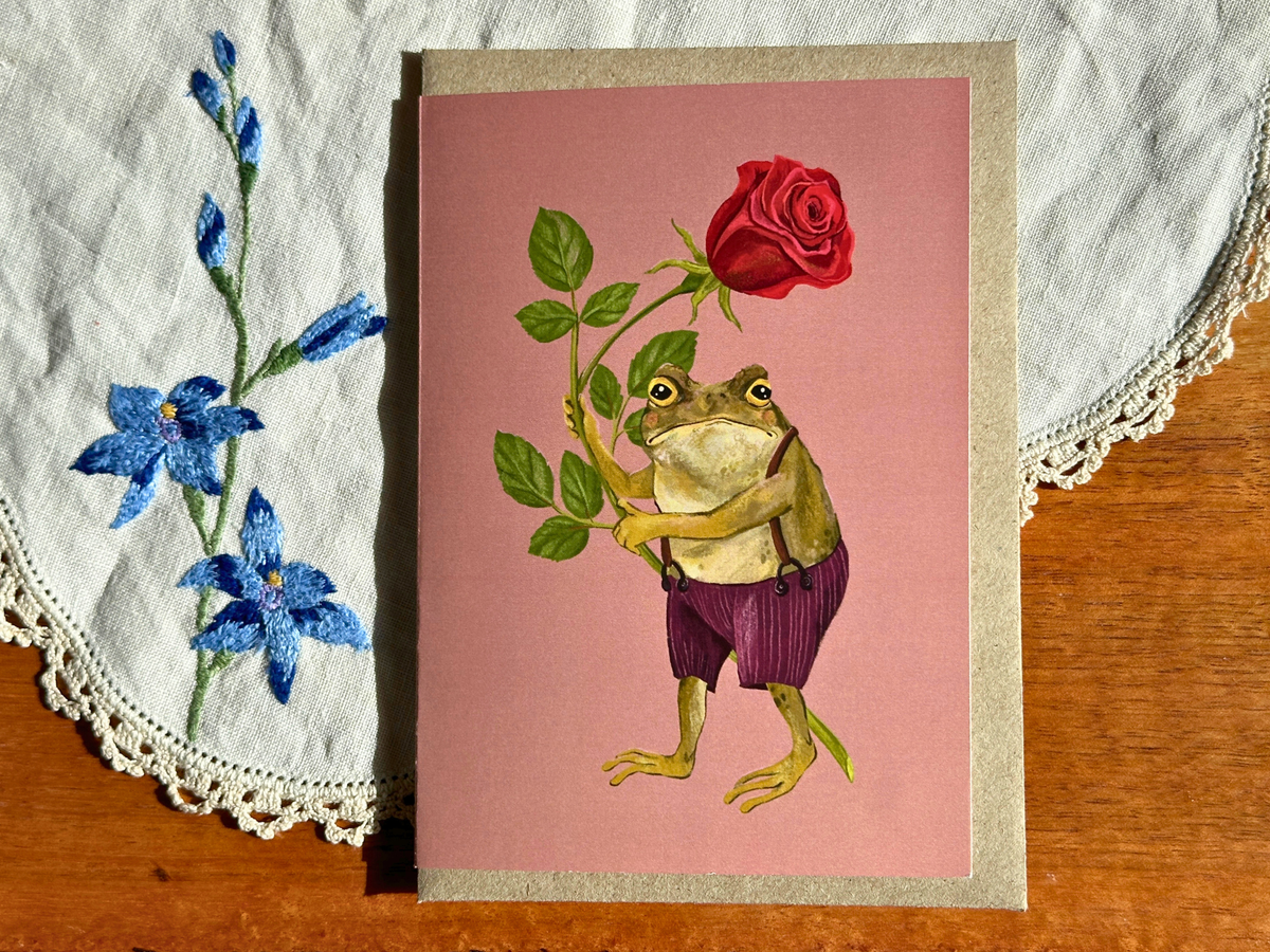 Love from Gustav greeting card with a toad offering a red rose, laid on an embroidered doily on a wooden table