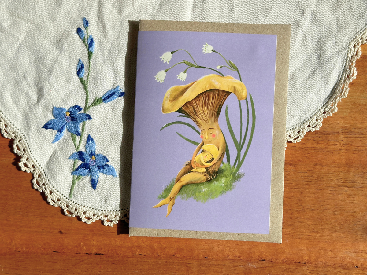 Mushroom Snuggles greeting card with a mushroom hugging its baby on lilac, on an embroidered doily on a wooden table