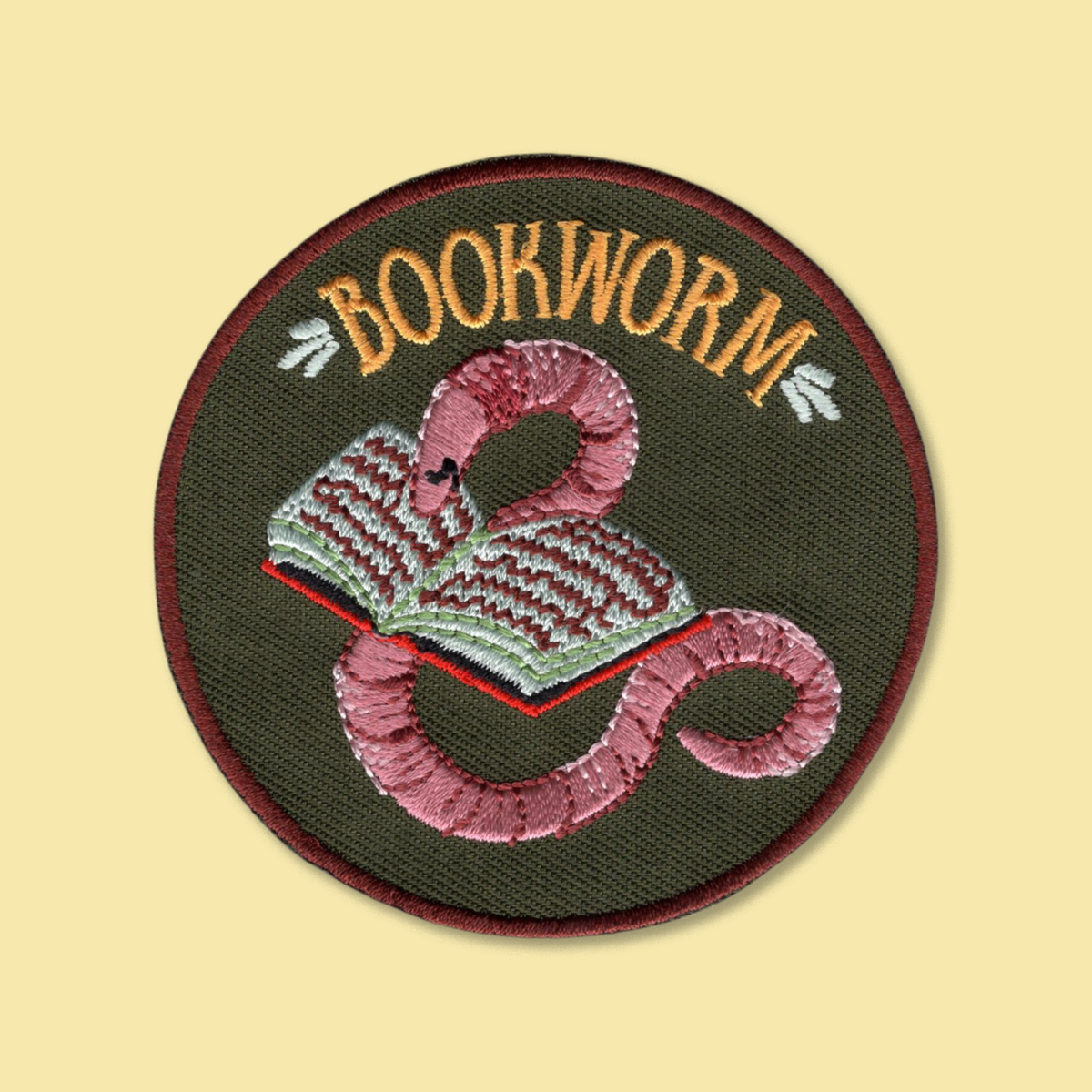 Bookworm iron-on patch by Anna Seed: a pink embroidered earthworm curled around an open book on dark green