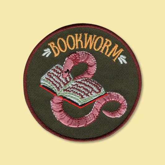 Bookworm iron-on patch by Anna Seed: a pink embroidered earthworm curled around an open book on dark green