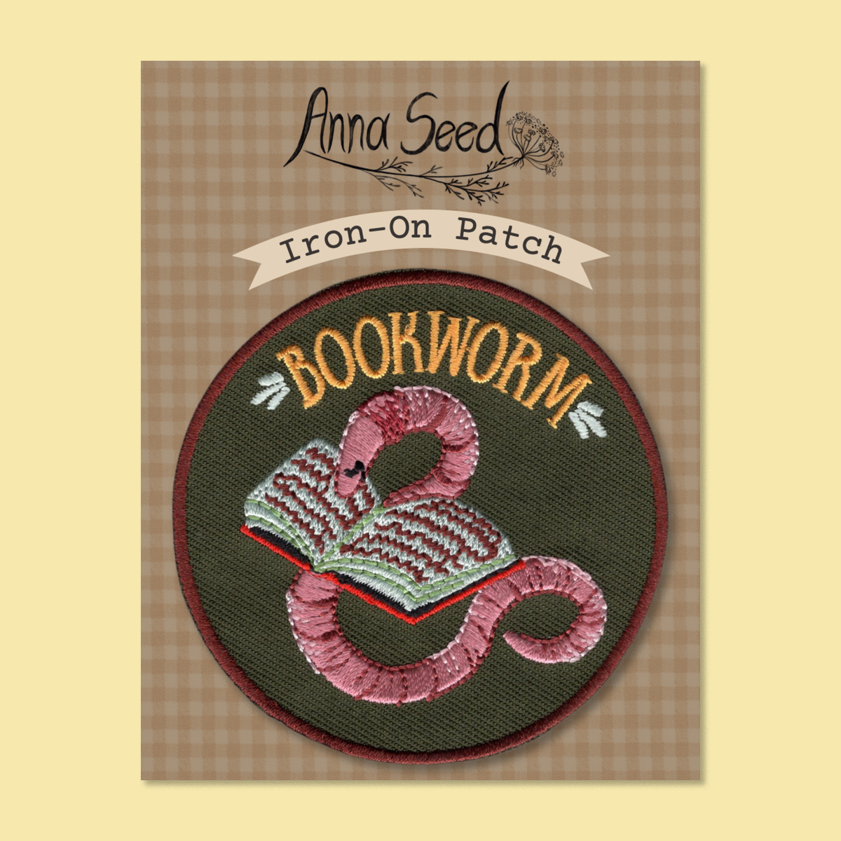 Bookworm patch on its gingham Iron-On Patch backing card, pink worm and open book stitched on a dark green circle
