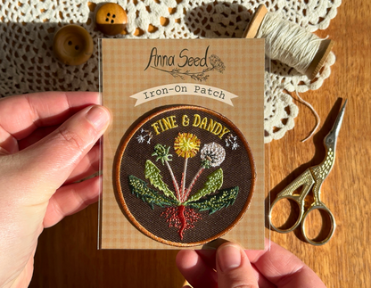 Fine & Dandy dandelion patch in its packaging held over a wooden table with lace doily, buttons, thread and scissors