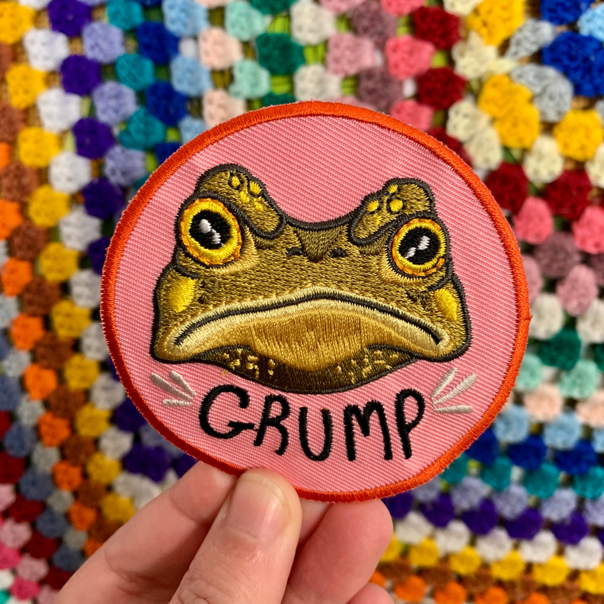 Gustav the Grumpy Toad patch held in a hand in front of a colourful crochet granny-square blanket