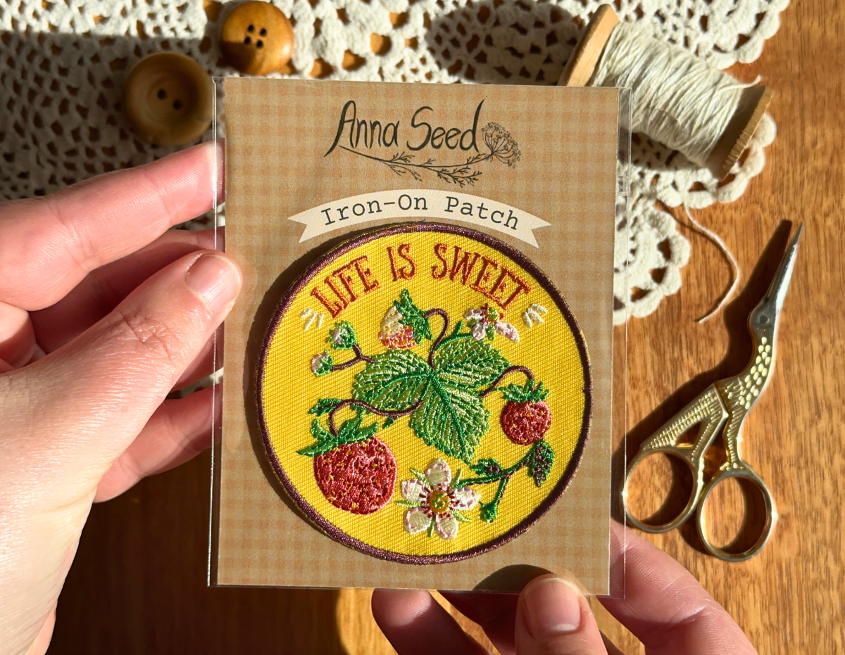 Life Is Sweet strawberry patch in its packaging held over a wooden table with lace doily, buttons, thread and scissors