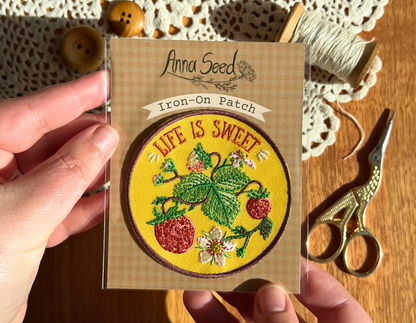 Life Is Sweet strawberry patch in its packaging held over a wooden table with lace doily, buttons, thread and scissors