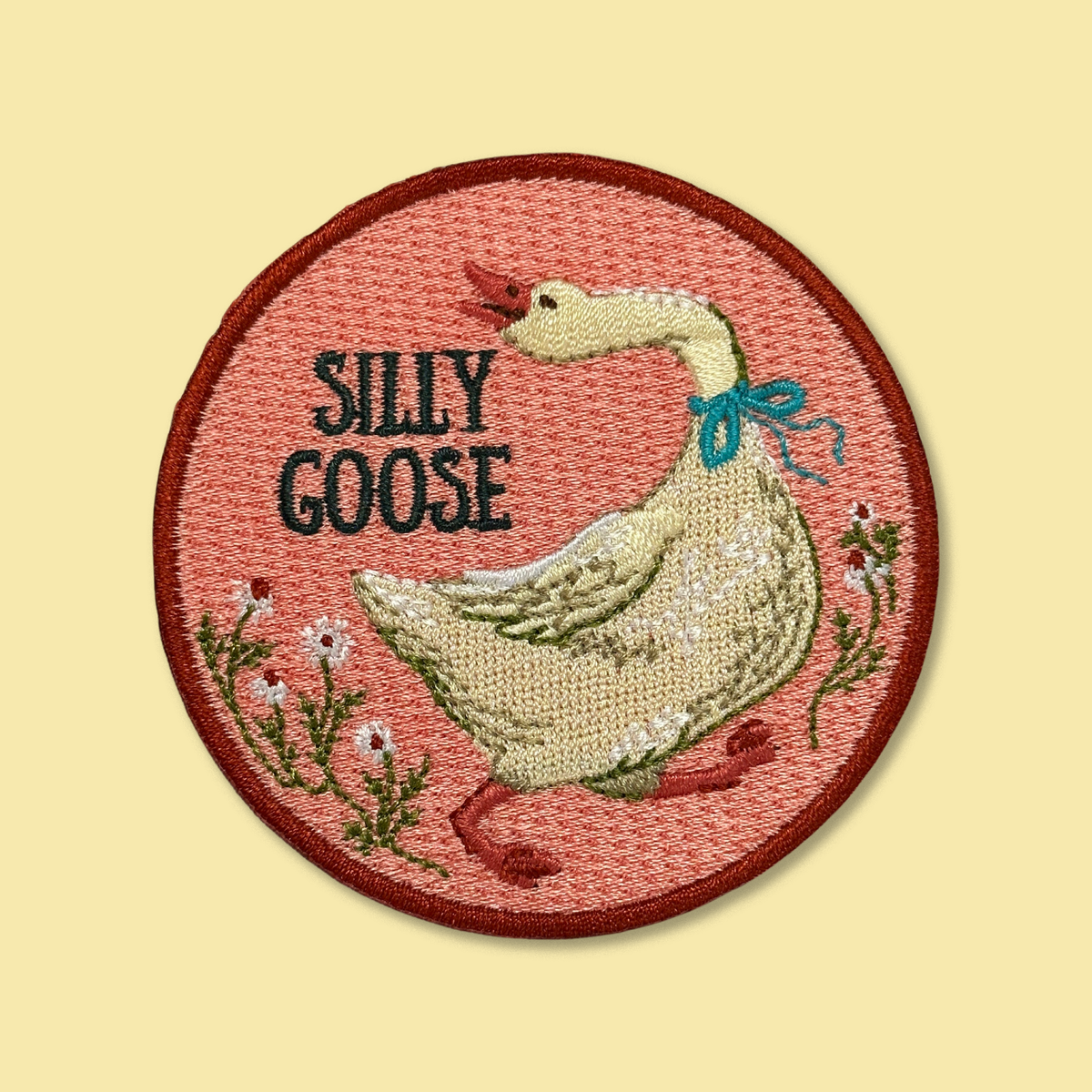 Silly Goose iron-on patch by Anna Seed: an embroidered white goose with a blue neck ribbon honking among daisies on pink