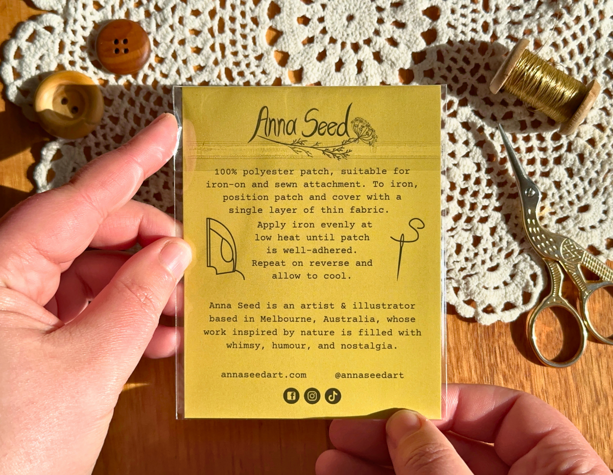 Back of a packaged iron-on patch card with ironing instructions and artist bio, held over lace and gold scissors