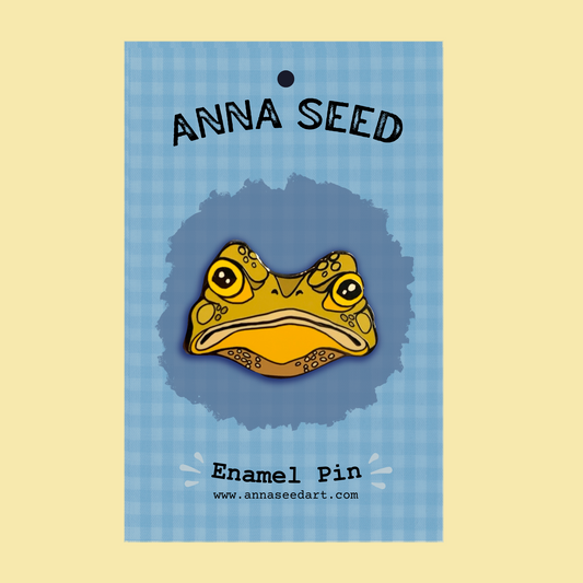 Gustav the Grumpy Toad enamel pin by Anna Seed, a frowning yellow toad face on a blue gingham backing card