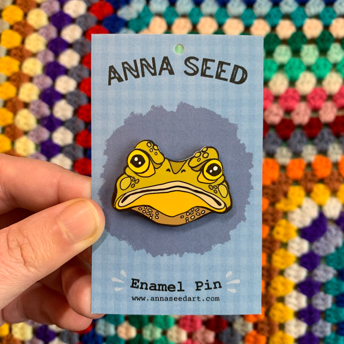 Gustav the Grumpy Toad enamel pin on its blue backing card, held in a hand in front of a colourful crochet blanket