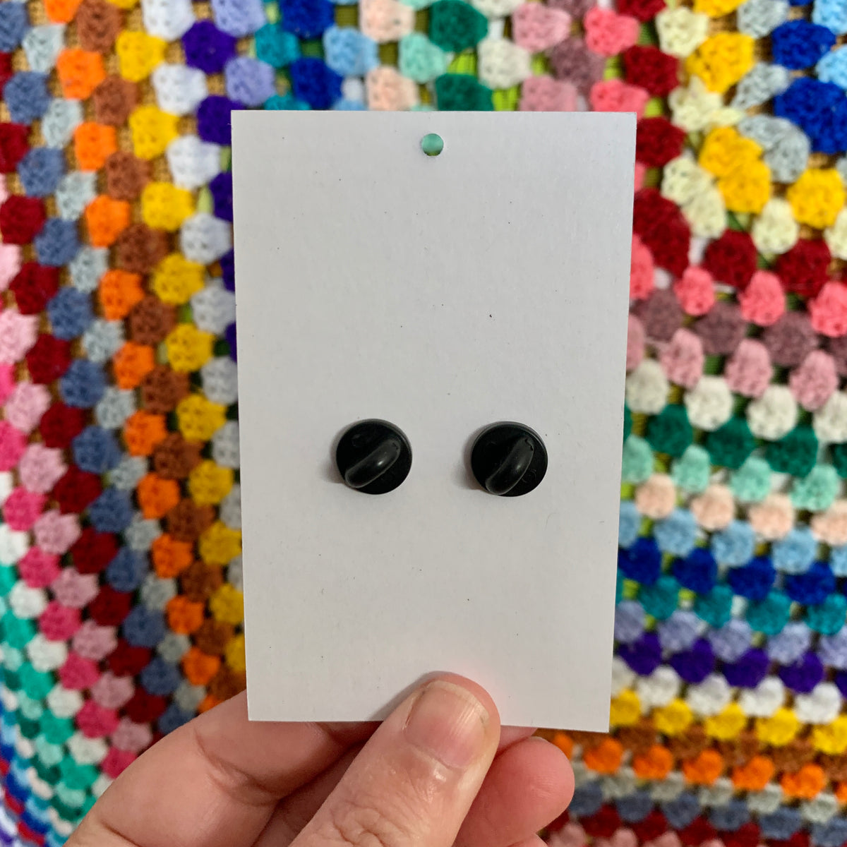 Back of the Gustav the Grumpy Toad pin card showing two black rubber clutch backs, held against a rainbow crochet blanket