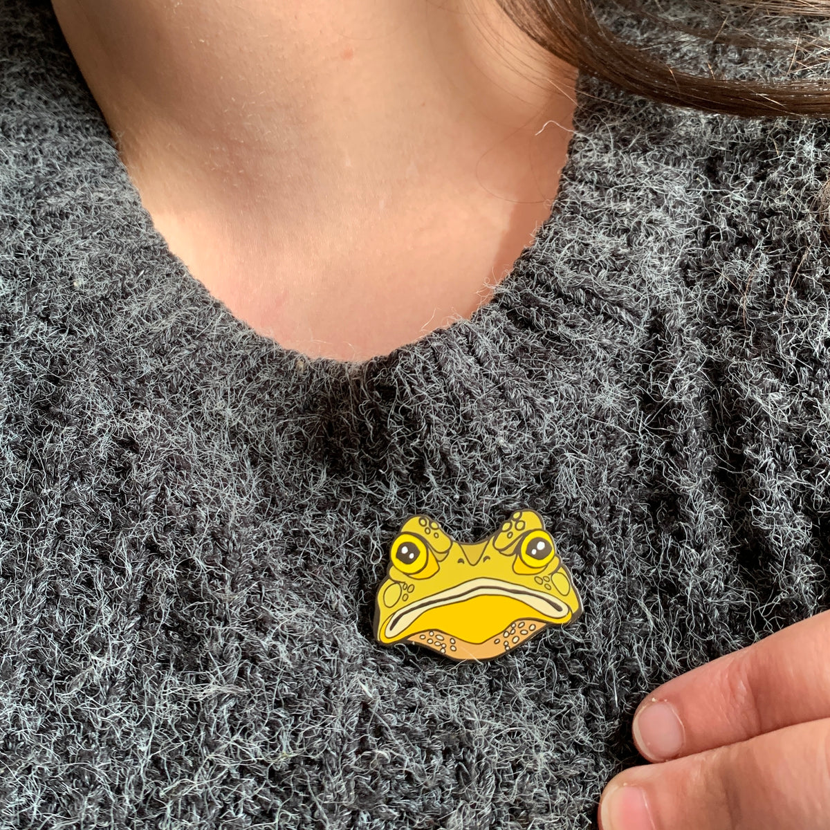 Gustav the Grumpy Toad enamel pin worn on a fluffy charcoal knit jumper, just below the neckline