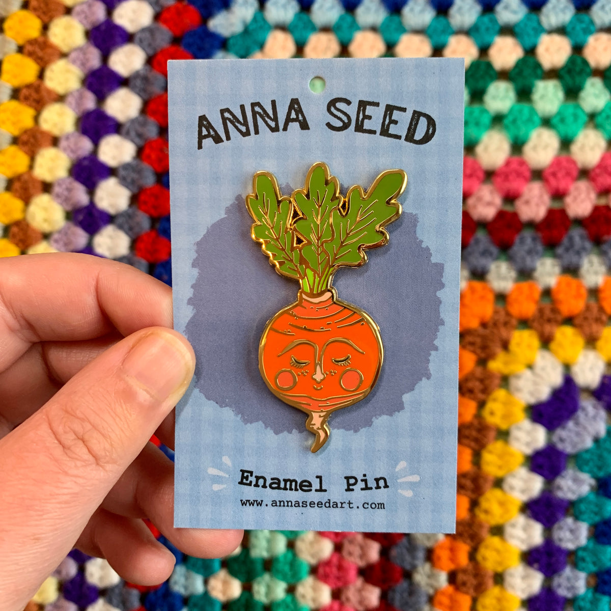 Sweet Radish enamel pin on its blue backing card, held in a hand in front of a colourful crochet blanket