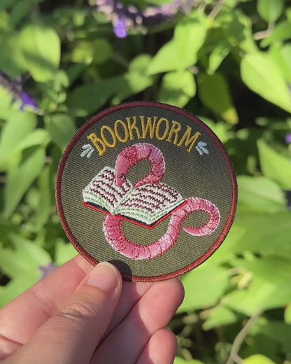 Video: the Bookworm patch held in a hand in front of sunlit green leaves, showing its pink worm and open book embroidery
