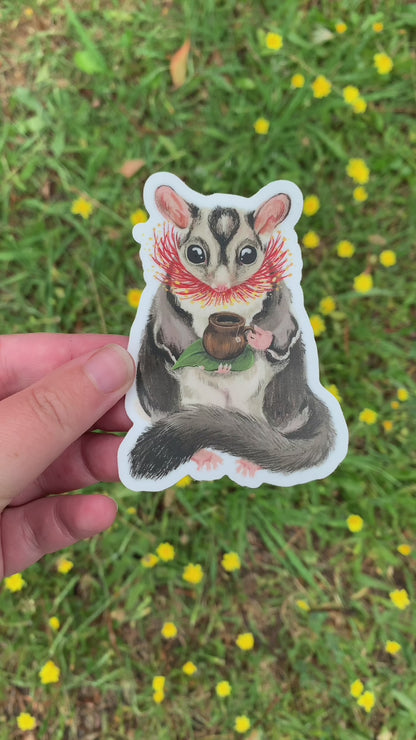 Video: the sugar glider teatime sticker held in a hand above green grass dotted with yellow flowers