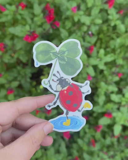 Video: the ladybird sticker with clover umbrella held in a hand in front of a green shrub with red flowers