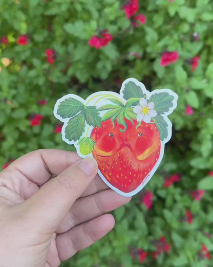 Video: the smiling strawberry sticker held in a hand in front of a green garden shrub with red flowers