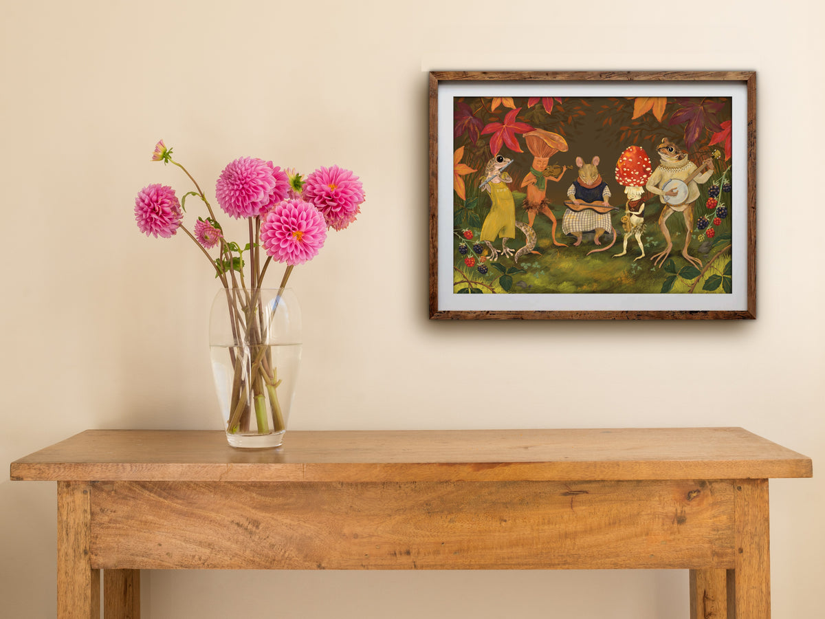 Autumn Jam landscape print of a woodland band, framed on a cream wall above a wooden table with pink dahlias