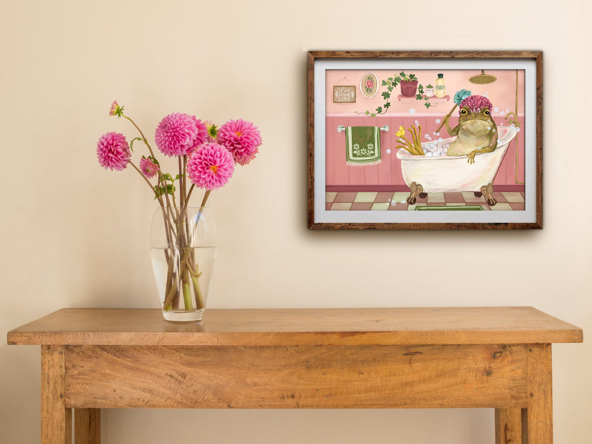 Bath Time print of a toad in a pink bathroom, framed on a cream wall above a wooden table with pink dahlias