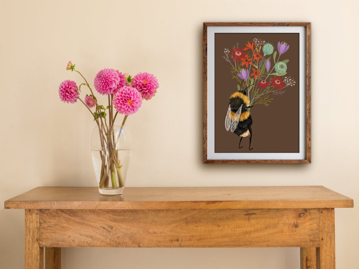 Bee with a Posy print of a bumblebee and its bouquet, framed on a cream wall above a table with pink dahlias