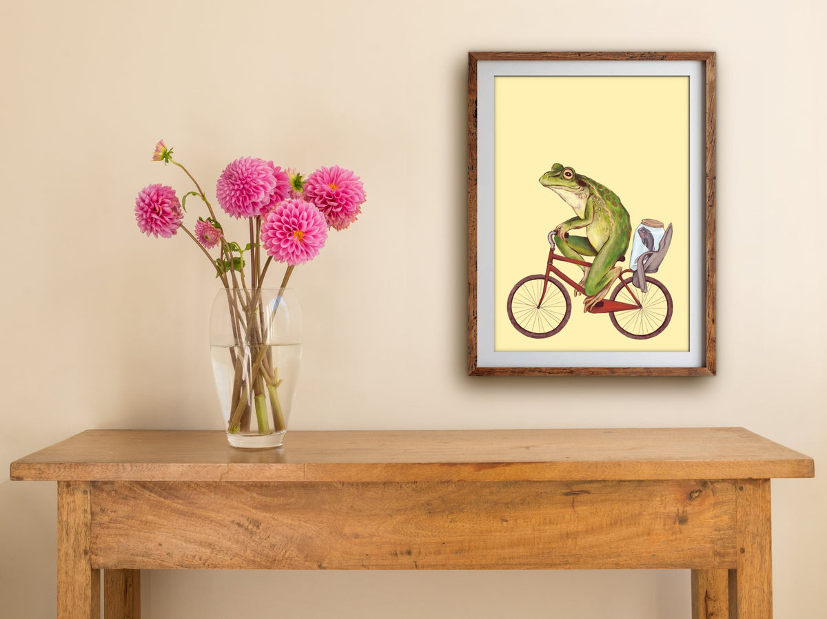 Bicycle Frog print of a frog on a red bicycle, framed on a cream wall above a wooden table with pink dahlias