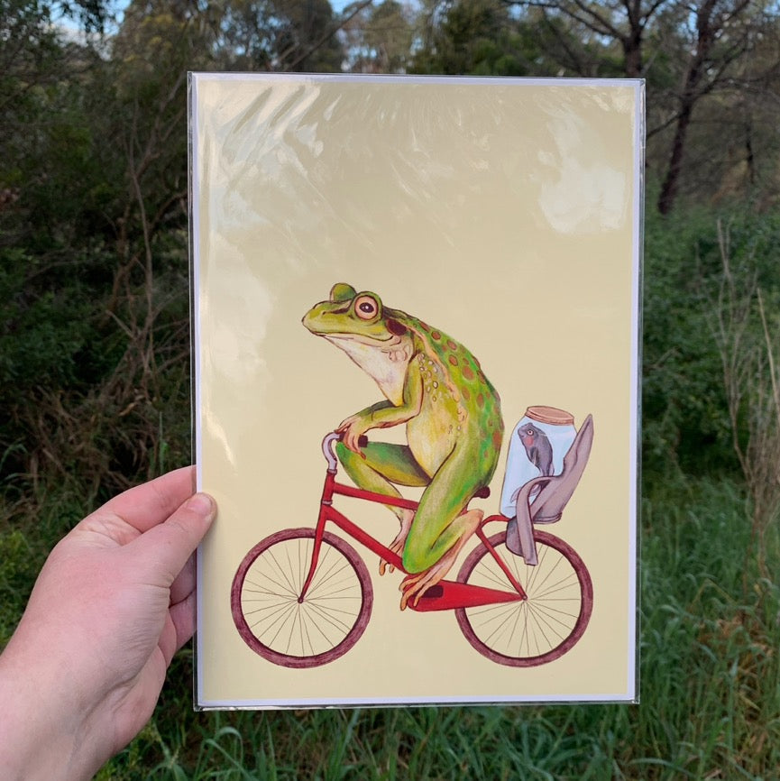 Hand holding the Bicycle Frog art print in a clear sleeve outdoors, in front of green bushland