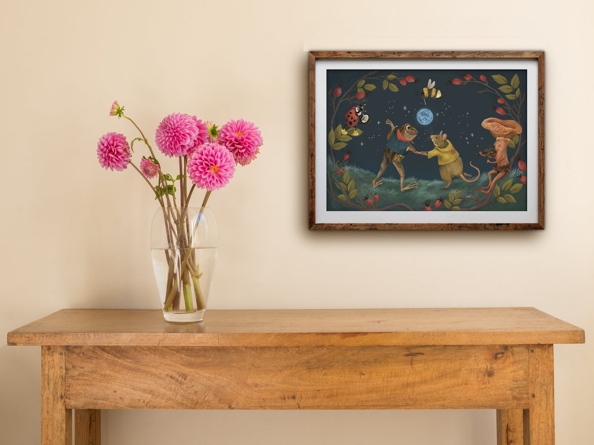 Blue Moon landscape print of a moonlit woodland dance, framed on a cream wall above a table with pink dahlias