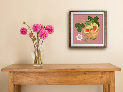 Blushing Berry print of a rosy-cheeked strawberry, framed on a cream wall above a wooden table with pink dahlias