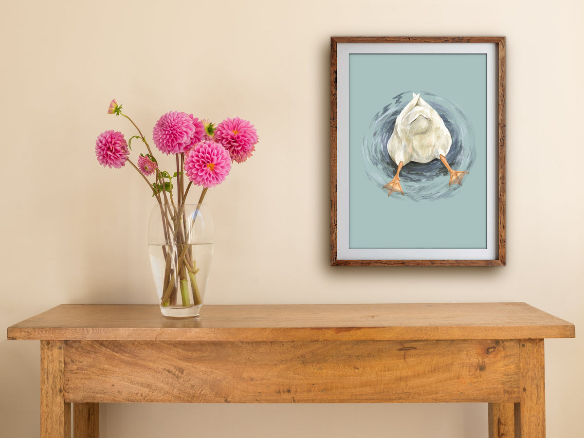 Framed Butt Quack print of an upended white duck hanging above a wooden side table with pink dahlias in a glass vase