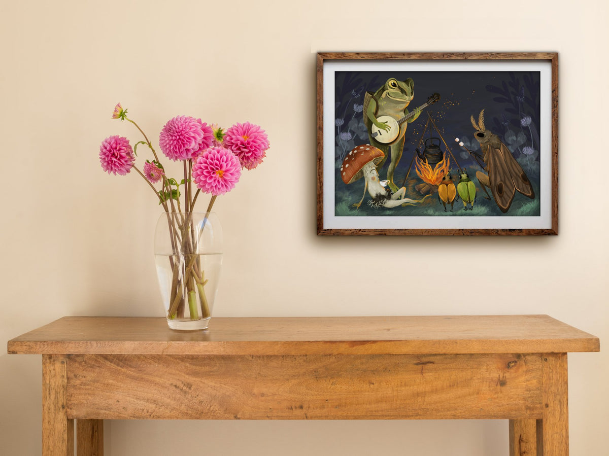 Framed Campfire print of a banjo-playing frog and woodland friends at night, hung above a wooden table with pink dahlias