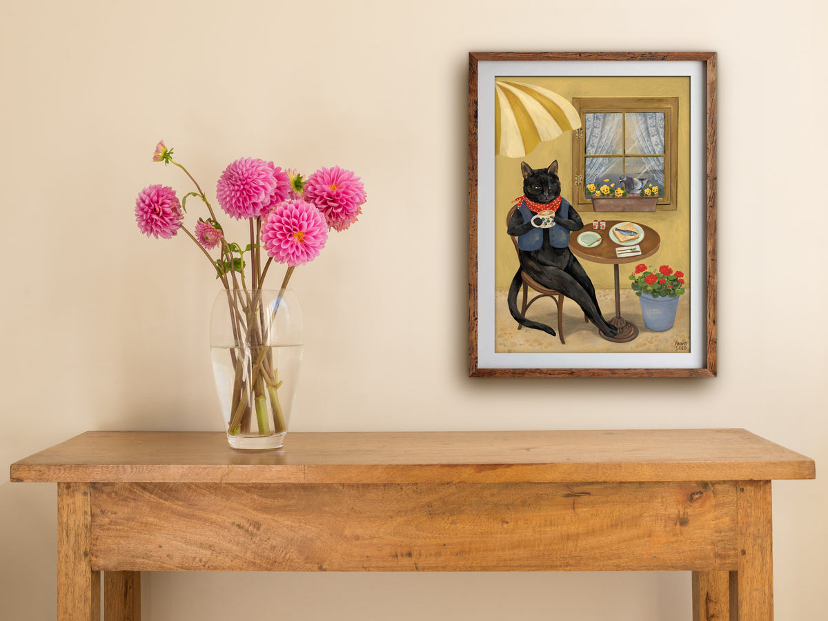 Framed Cat Café print of a black cat having coffee at an outdoor café table, hung above a wooden table with pink dahlias