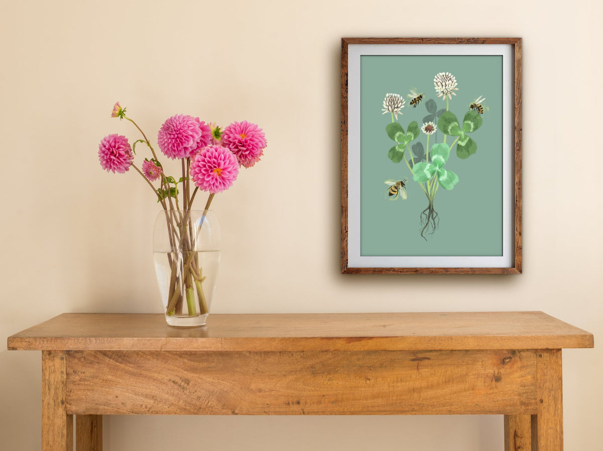 Framed Clover print of white clover and honeybees on sage green, hung above a wooden side table with pink dahlias in a vase