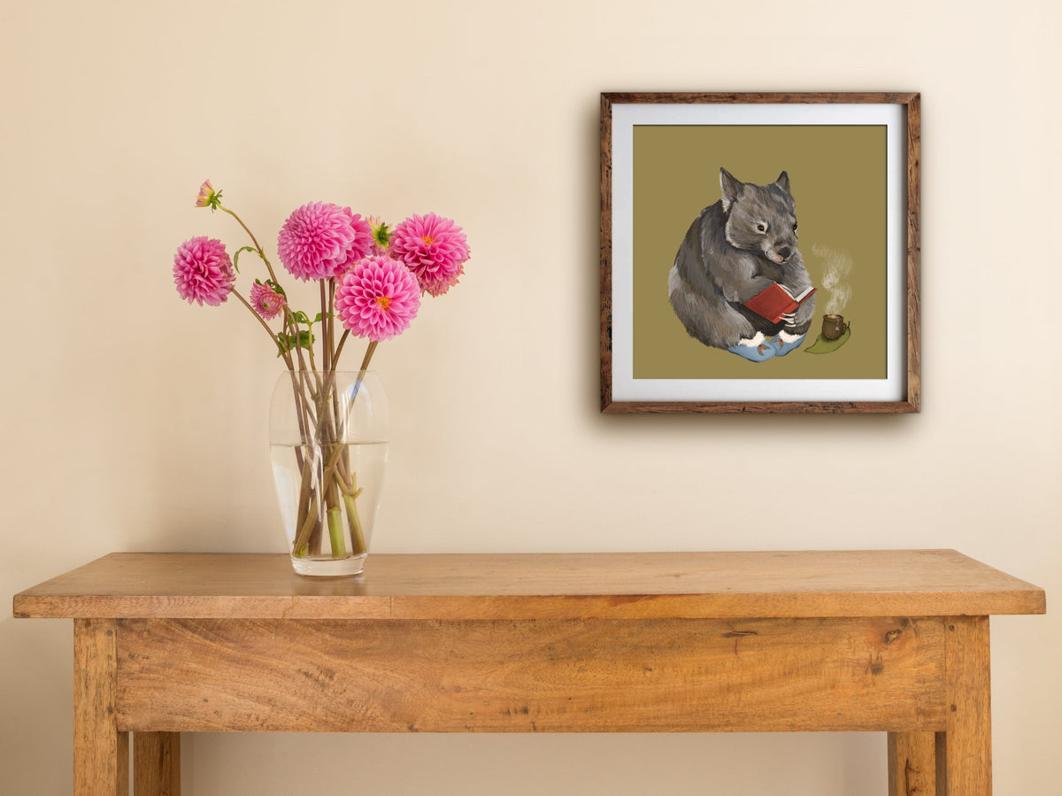 Framed Cosy Wombat print of a slipper-wearing wombat reading with tea, hung above a wooden side table with pink dahlias