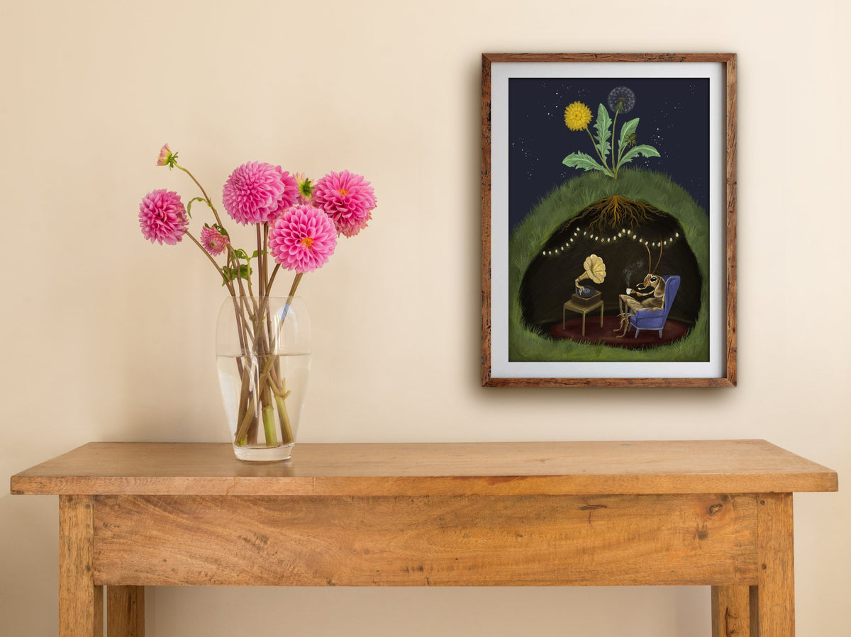 Framed Cricket at Home print of a cricket relaxing in its burrow beneath a dandelion, hung above a wooden table with dahlias
