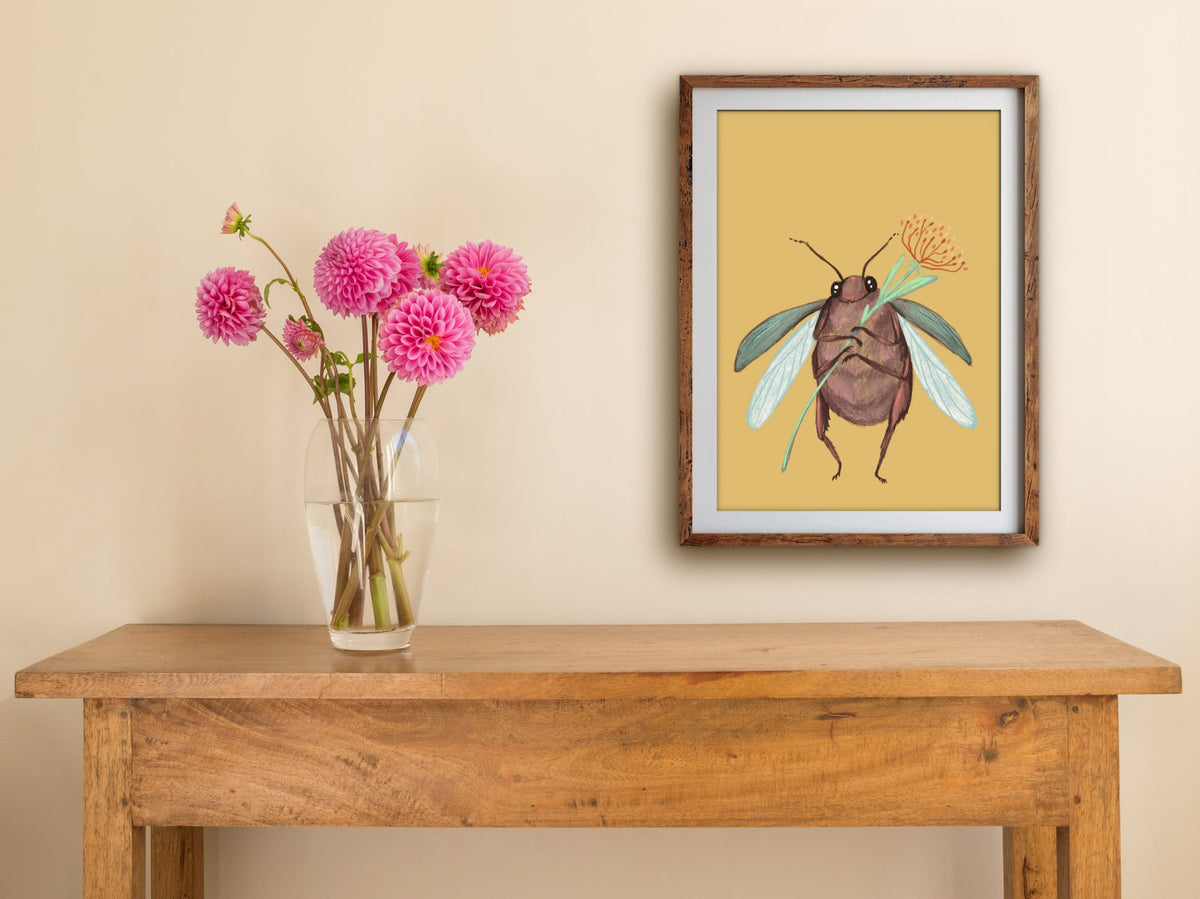 Framed Cute Beetle print of a beetle holding a flower on mustard yellow, hung above a wooden side table with pink dahlias