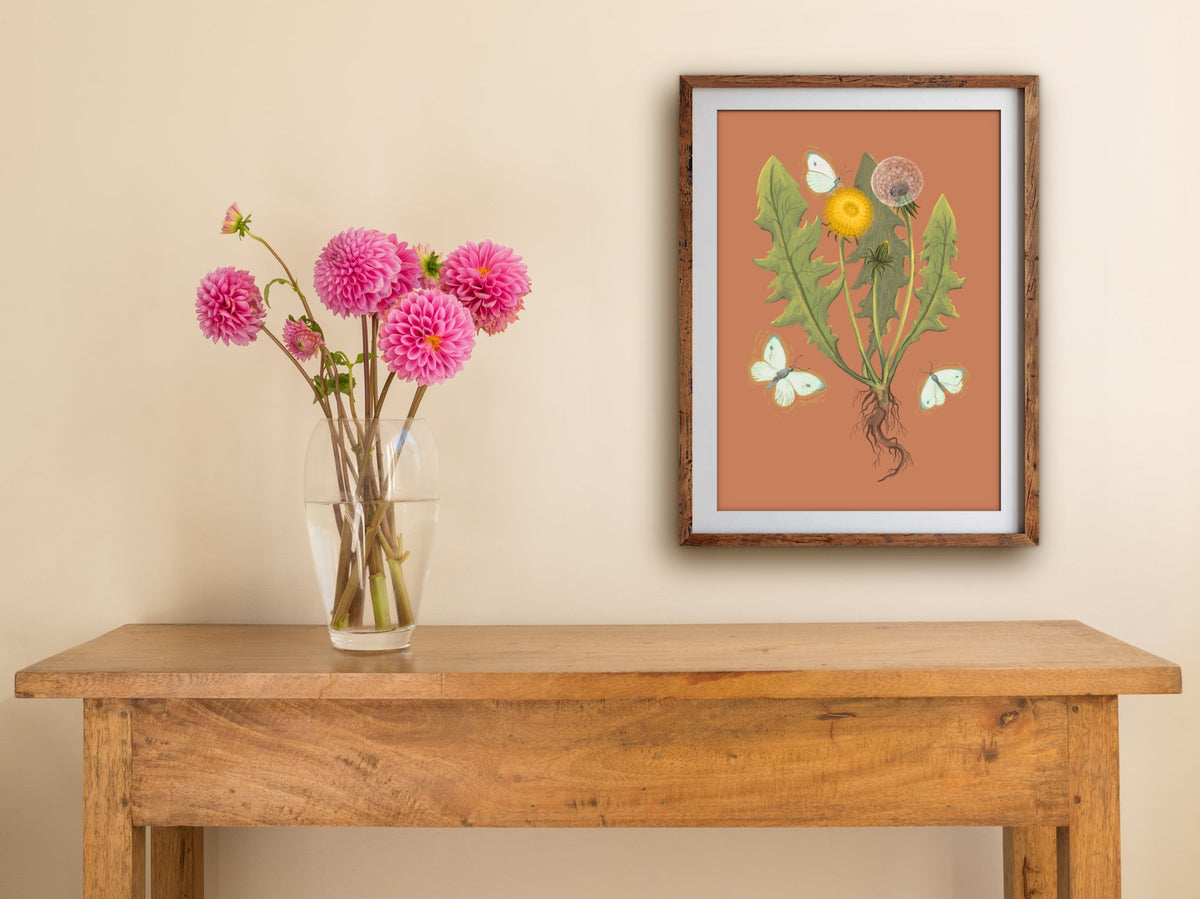 Framed Dandelion print of a dandelion and cabbage moths on burnt orange, hung above a wooden side table with pink dahlias