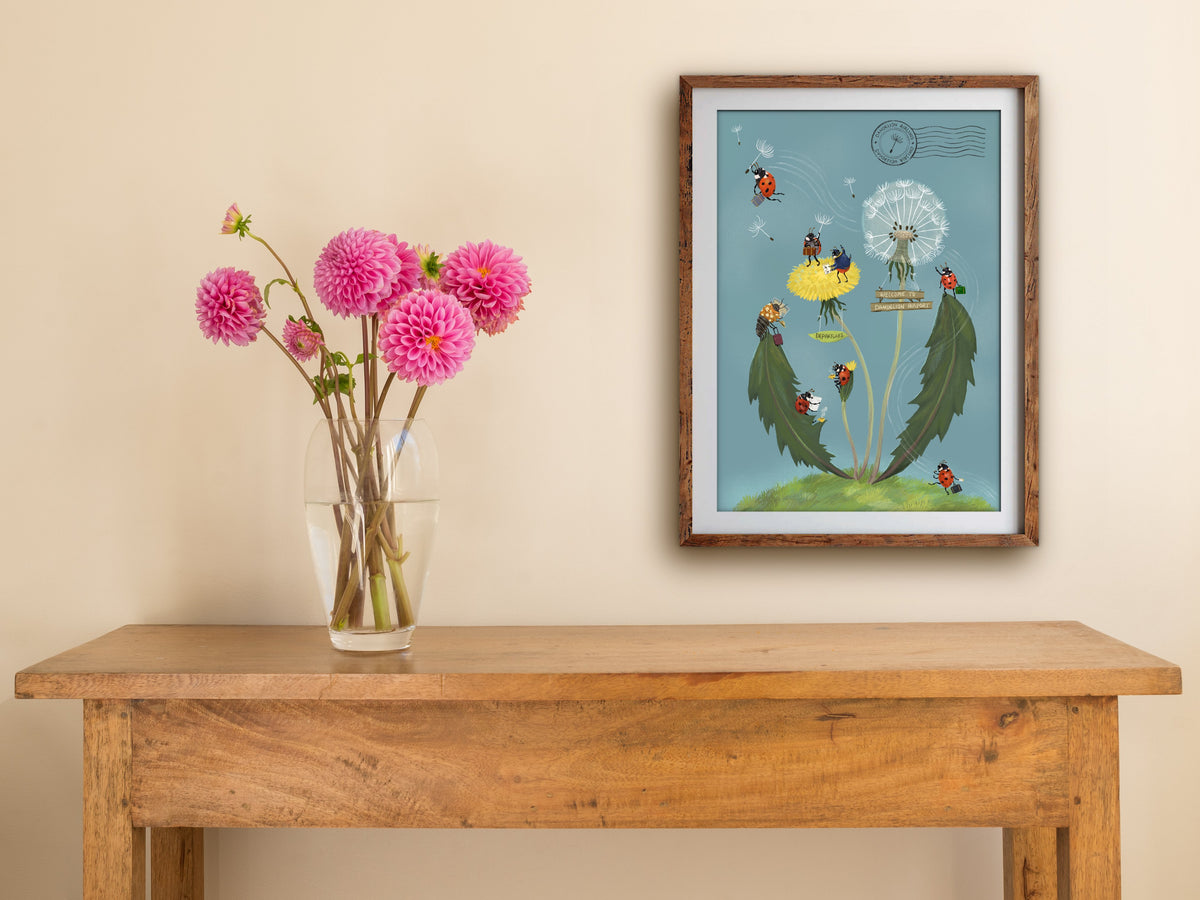 Framed Dandelion Airport print of ladybirds catching dandelion seed flights, hung above a wooden side table with pink dahlias